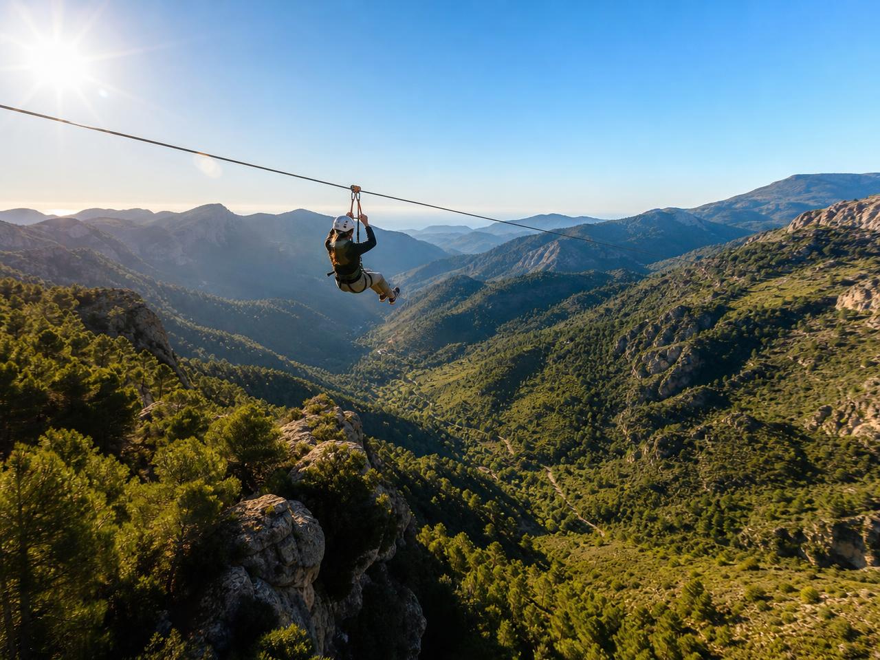 Person riding a long zipline high above a green Andalusian mountain valley under a bright blue sky